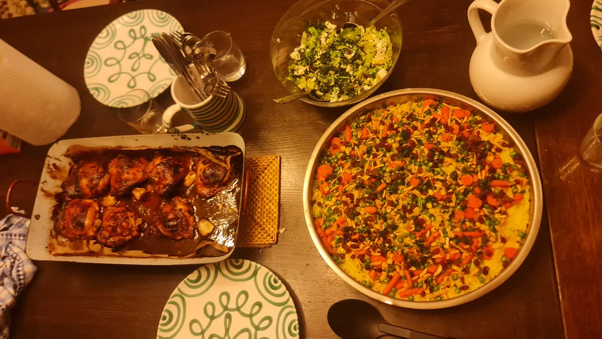 Evening dinner on a wooden table: a pan of rice with vegetables, a tray of baked chicken, salad and a water jug.