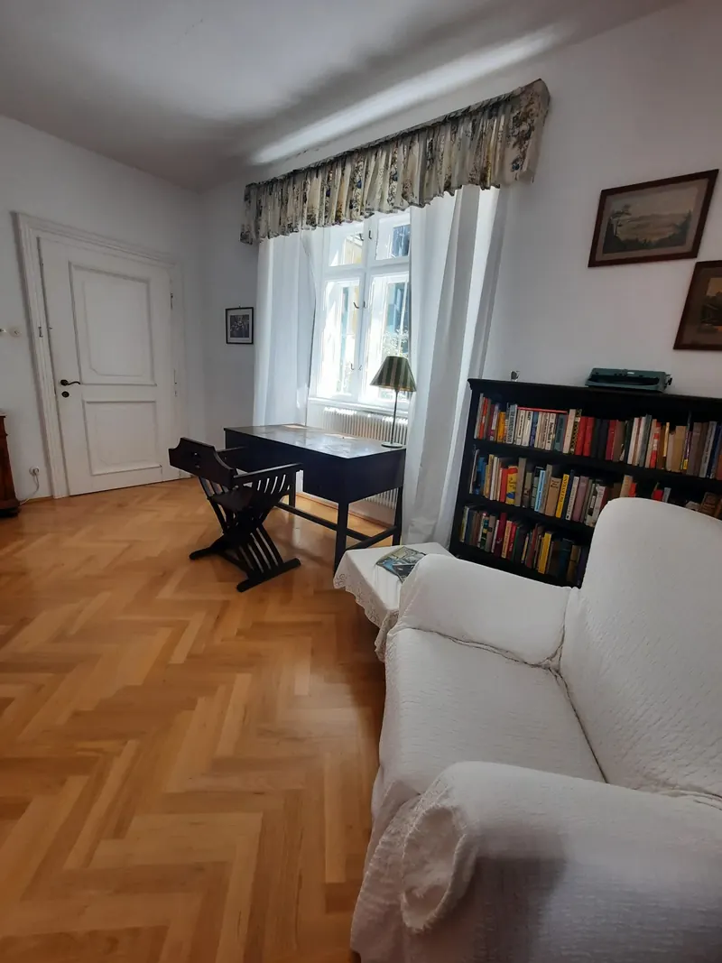 A writing table and folding chair by a window, next to shelves of books and a white armchair.