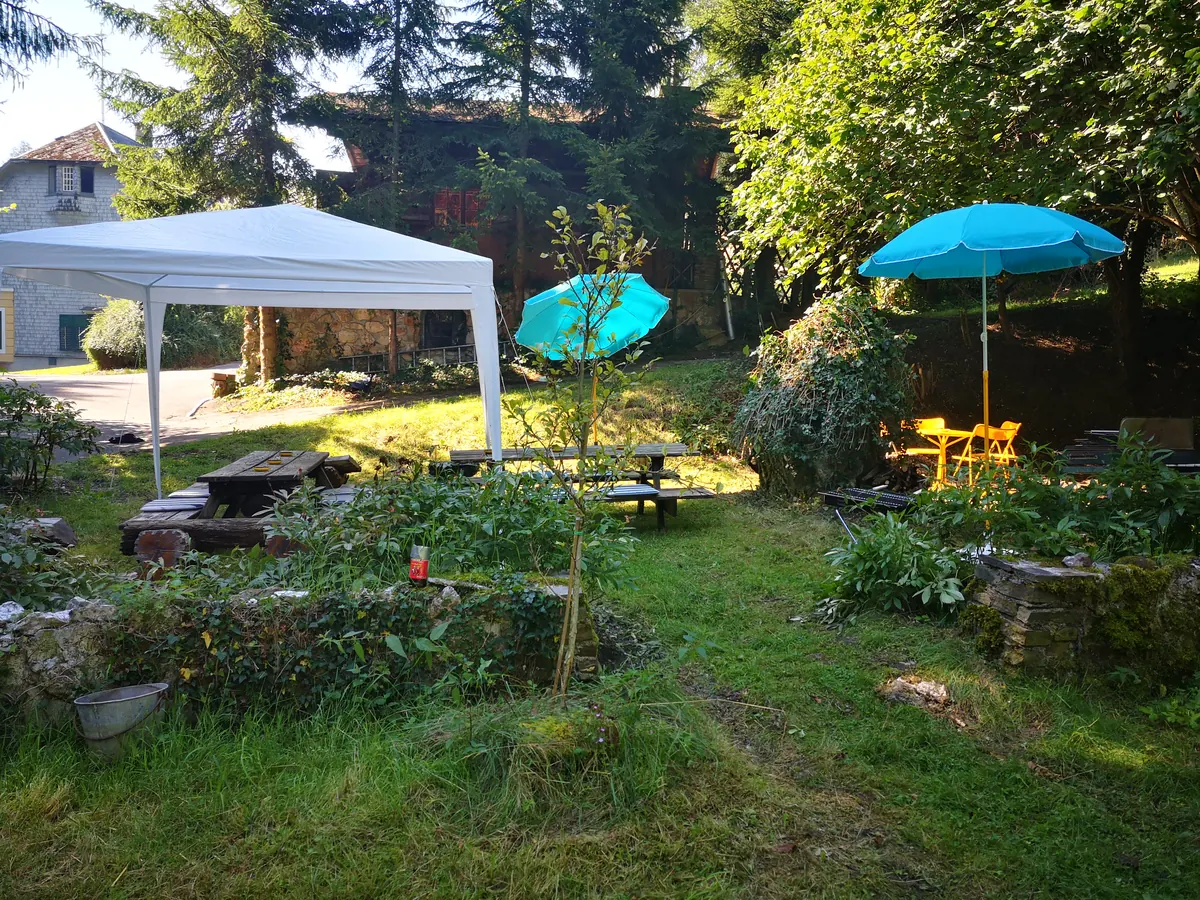 The garden in morning light: picnic tables, a white gazebo, two turquoise umbrellas and old trees in front of a stone house.